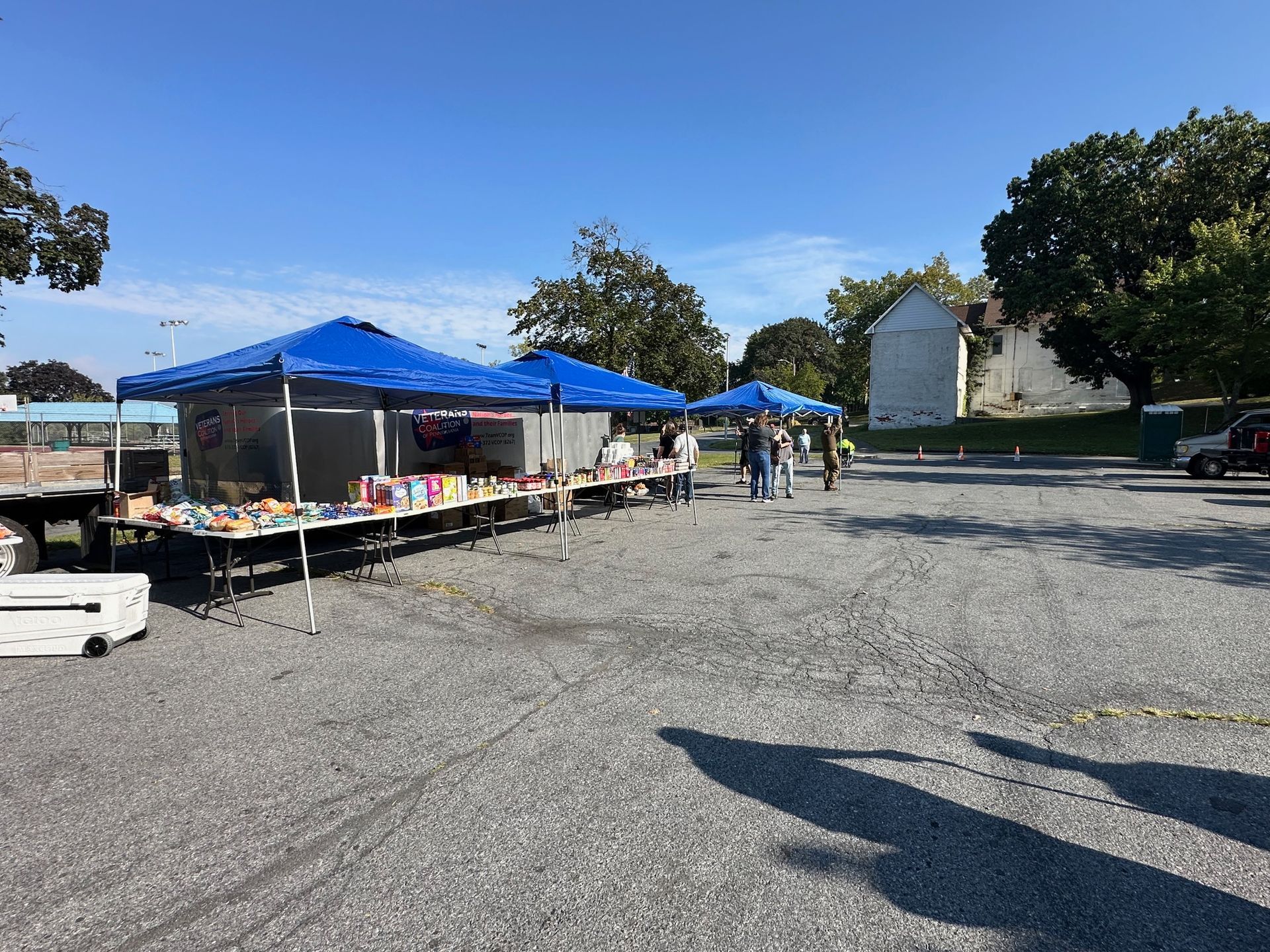 Outdoor market with blue canopy tents and tables on a gravel lot. People browse merchandise under a sunny, clear sky.