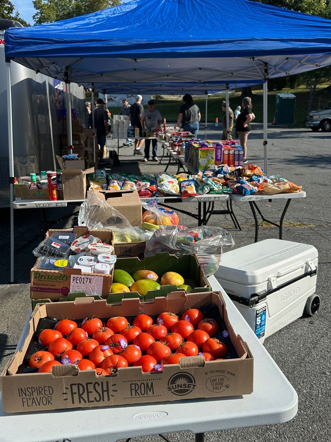 Farmers market stand with fresh produce and snacks under a blue tent. People browse on a sunny day.