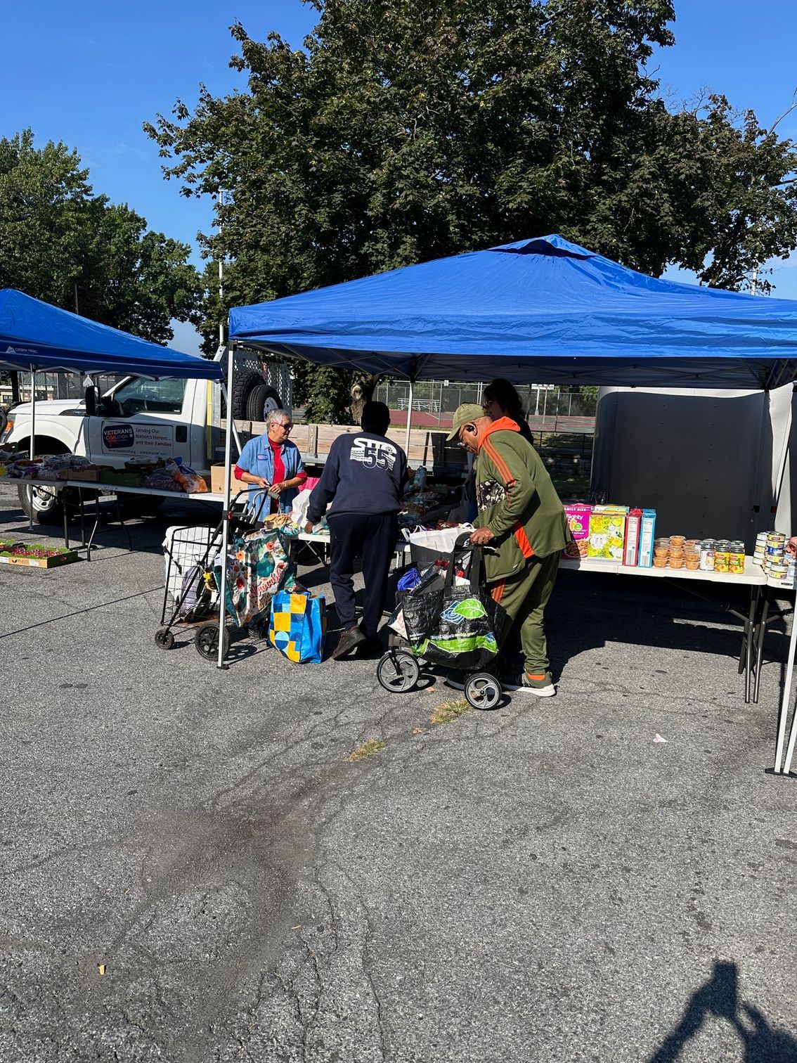 Outdoor market with blue canopy, people browsing items on tables. Sunny day.