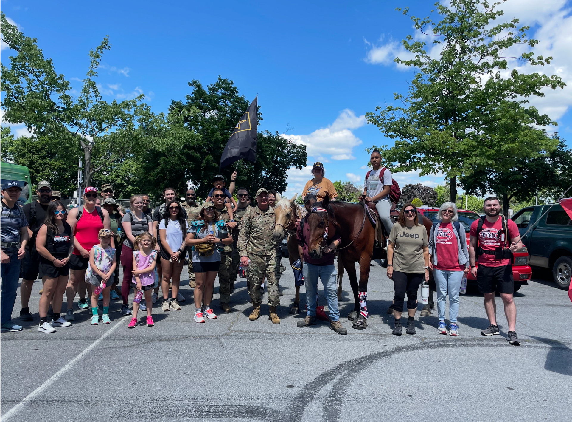Group of people with horses in a parking lot on a sunny day. Some are holding a flag.