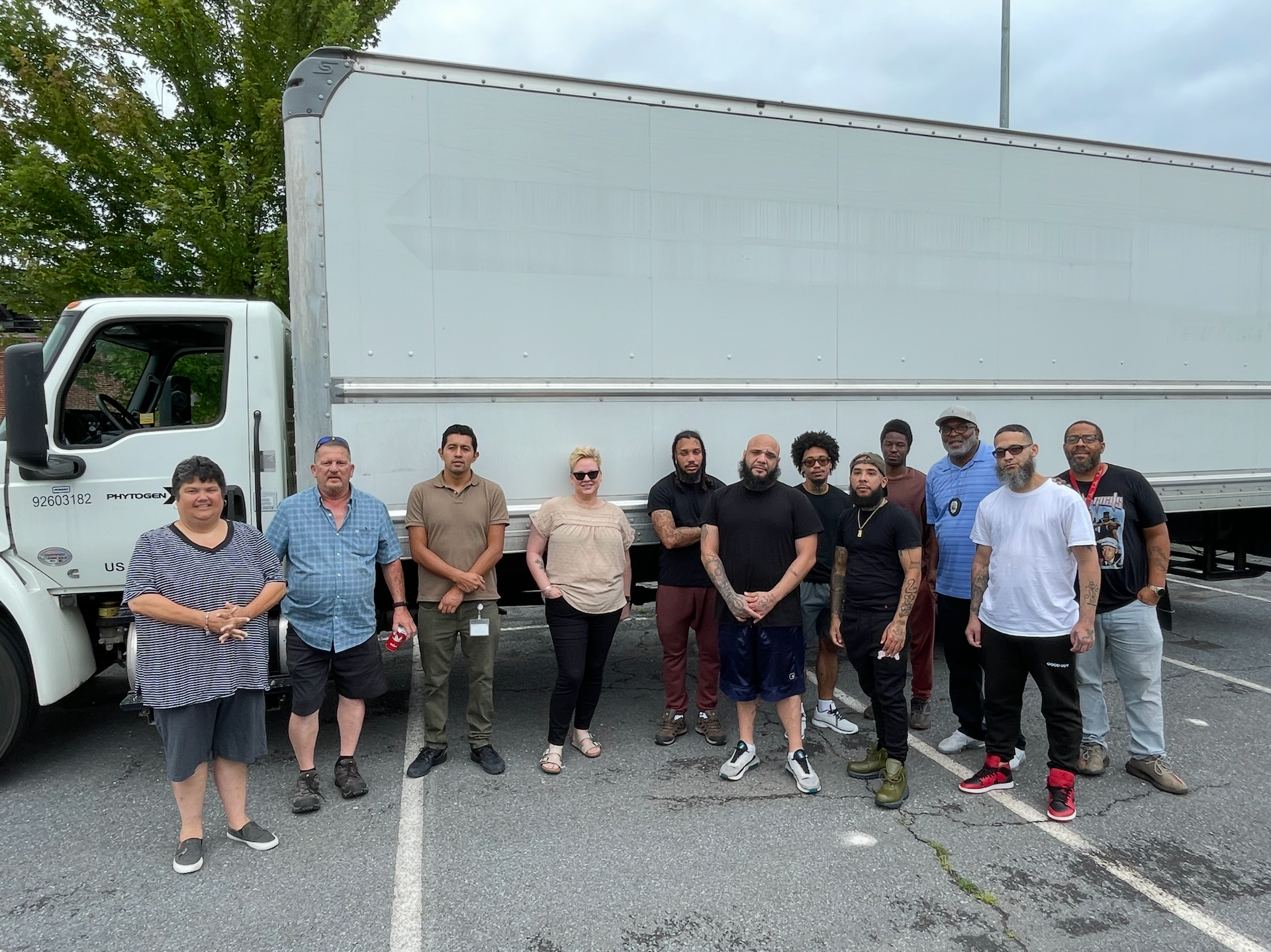 Group of people standing in front of a white truck. They are outside on a gray paved lot, overcast day.