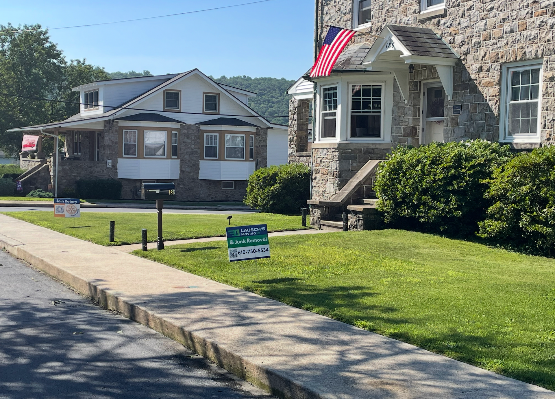 Stone houses with green lawns on a sunny day. American flag displayed, sidewalk in foreground.