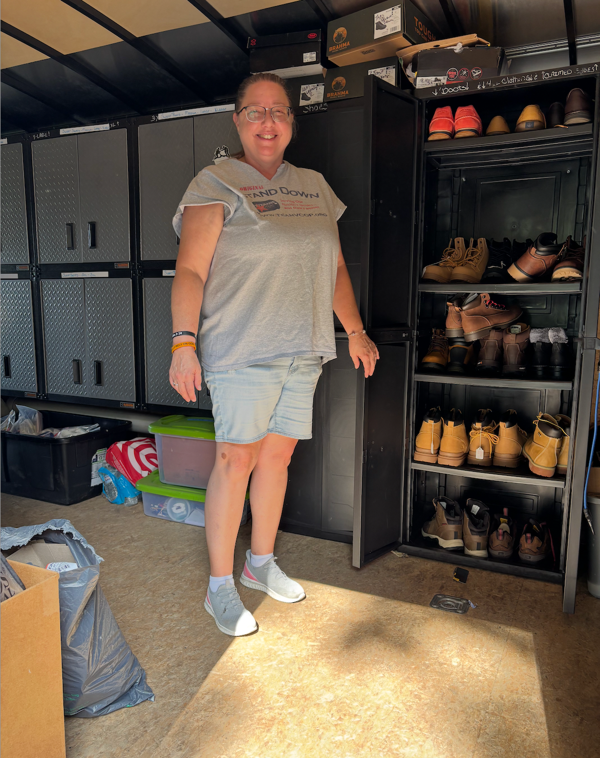 Woman standing near shoe organizer inside trailer, smiling. Gray hoodie, shorts. Shoes on shelves.