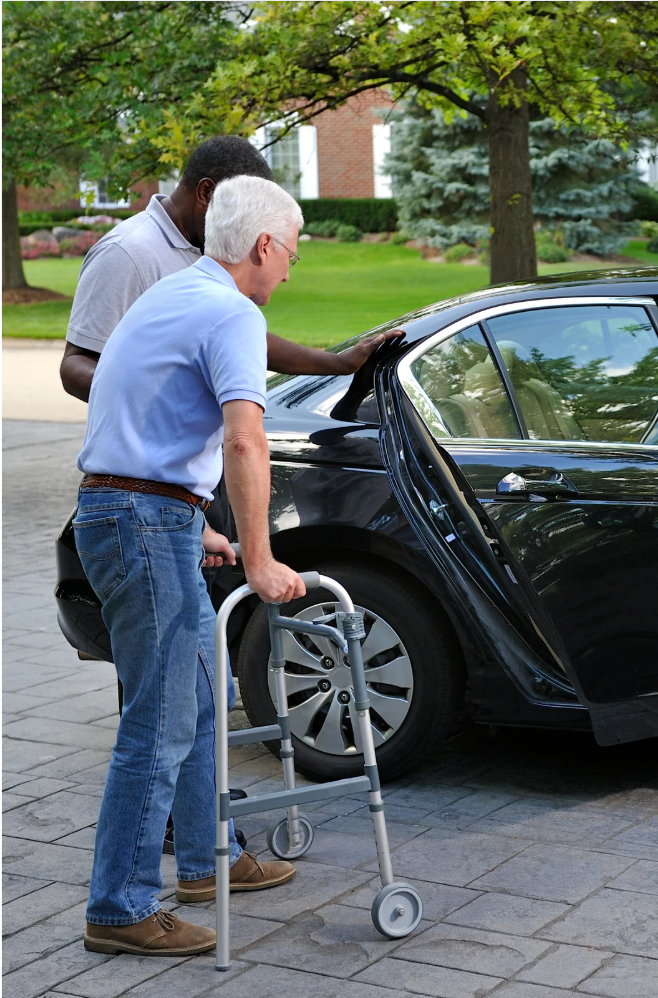 Man with walker assisted into a black car by another person. Outdoors, on a brick driveway.