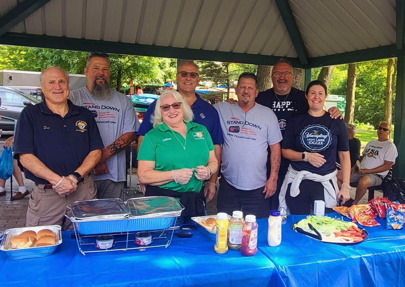 Group of people at an outdoor gathering, serving food from a table covered in blue cloth, under a shelter.