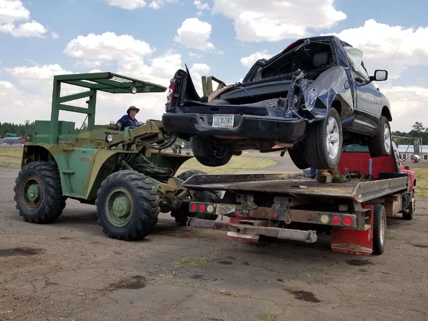 A green forklift lifts a heavily damaged black pickup truck onto the bed of a flatbed tow truck on a dirt lot.