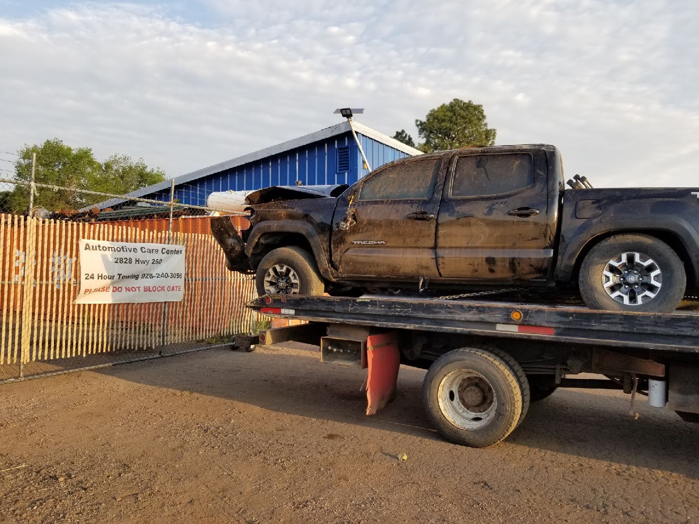 A damaged black pickup truck sits on the back of a flatbed tow truck in front of a bamboo fence and a blue building.