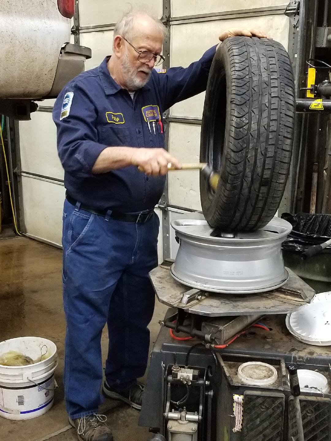 A mechanic in a blue uniform applies sealant to the inside of a car tire mounted on a rim in an auto shop.
