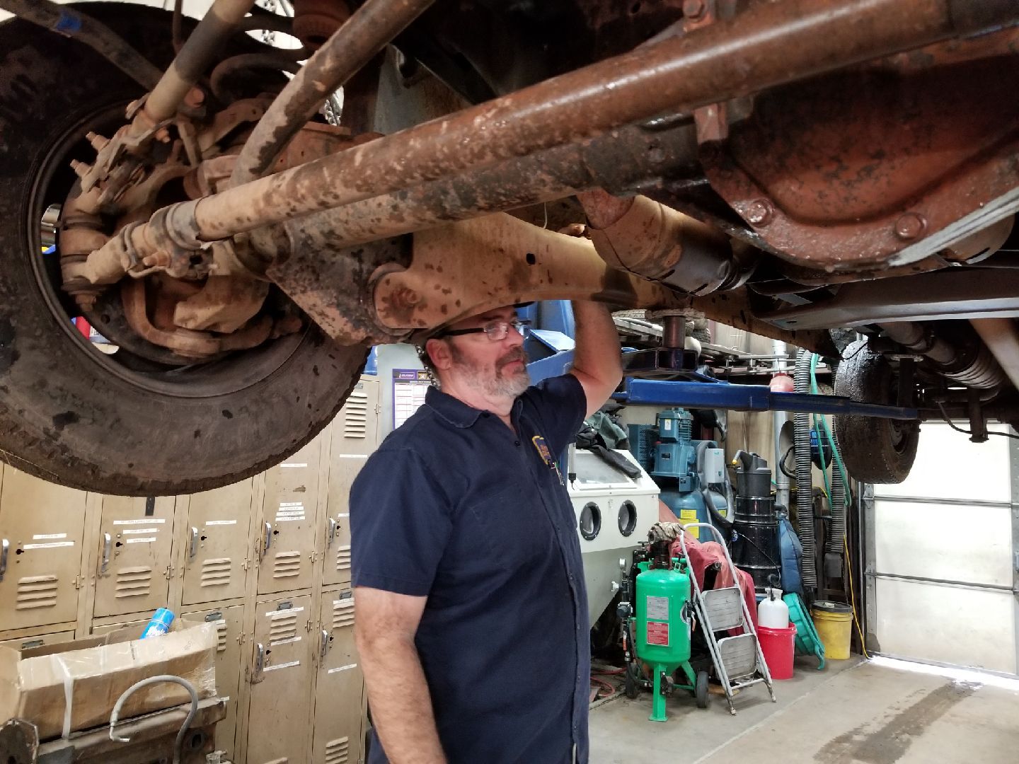A mechanic in a navy uniform inspects the undercarriage of a raised vehicle in a cluttered repair shop.