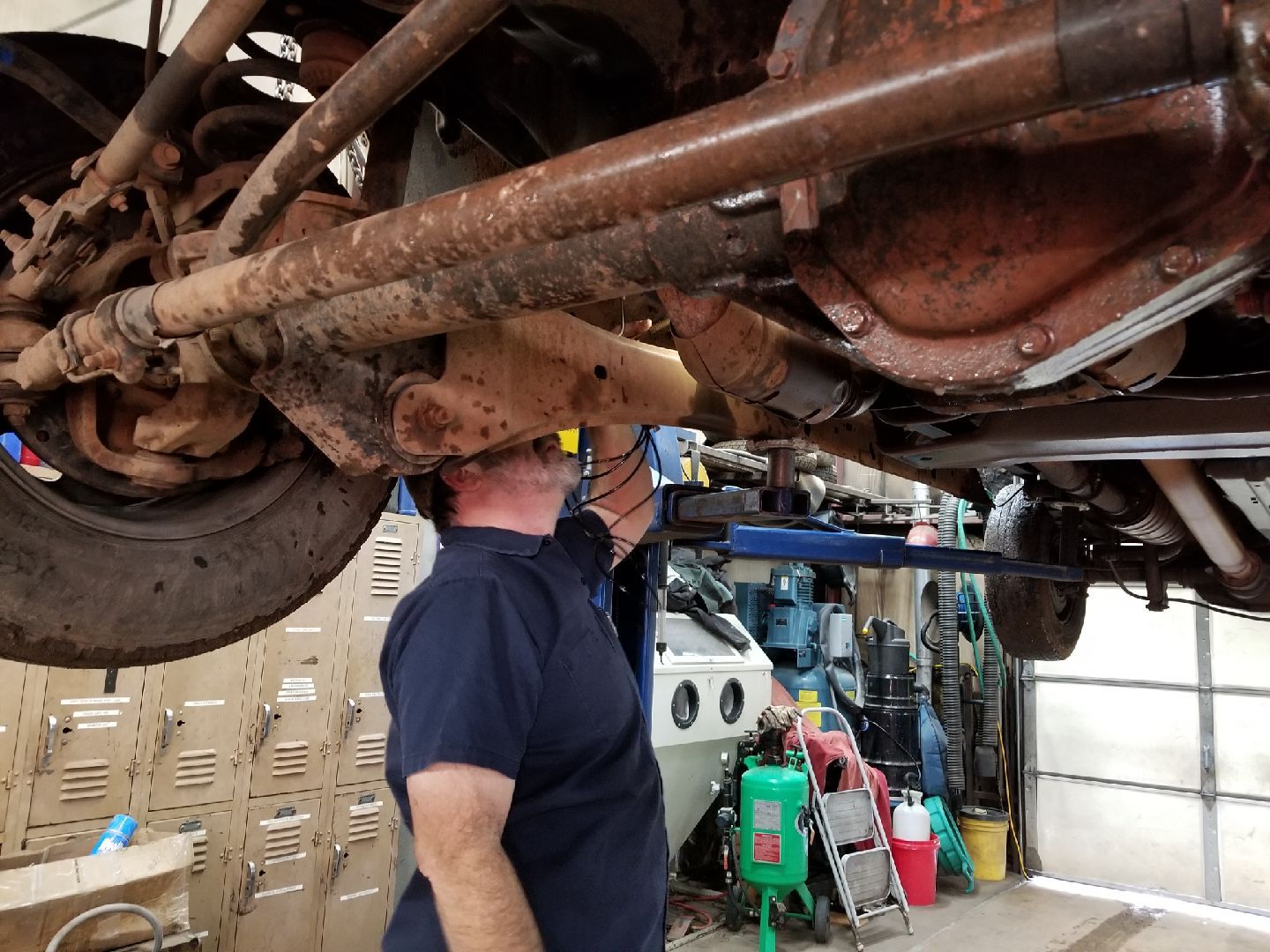 A mechanic stands under a vehicle hoisted on a lift, examining the rusted underside of the chassis and suspension.