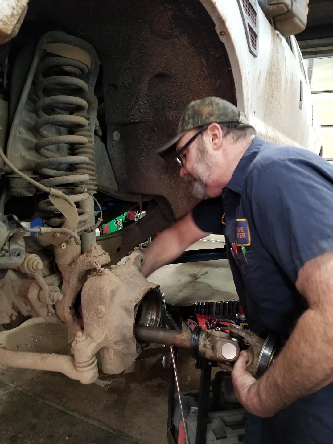 A mechanic in a garage works on the front suspension and axle assembly of a vehicle.