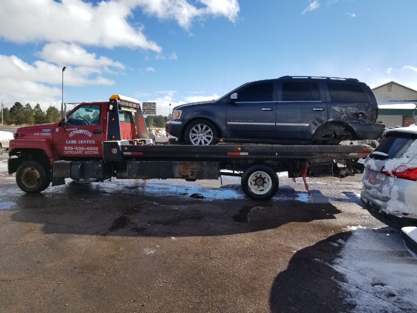 A red flatbed tow truck carries a damaged dark SUV through a snowy, outdoor lot.