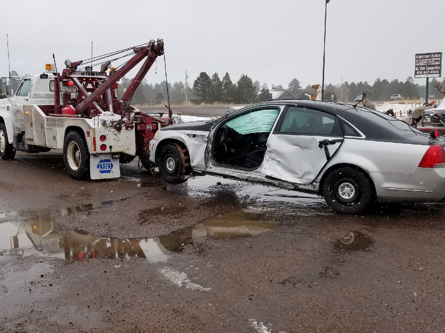 A tow truck hooks a heavily damaged silver sedan with a crushed passenger door and deployed airbags in a muddy lot.