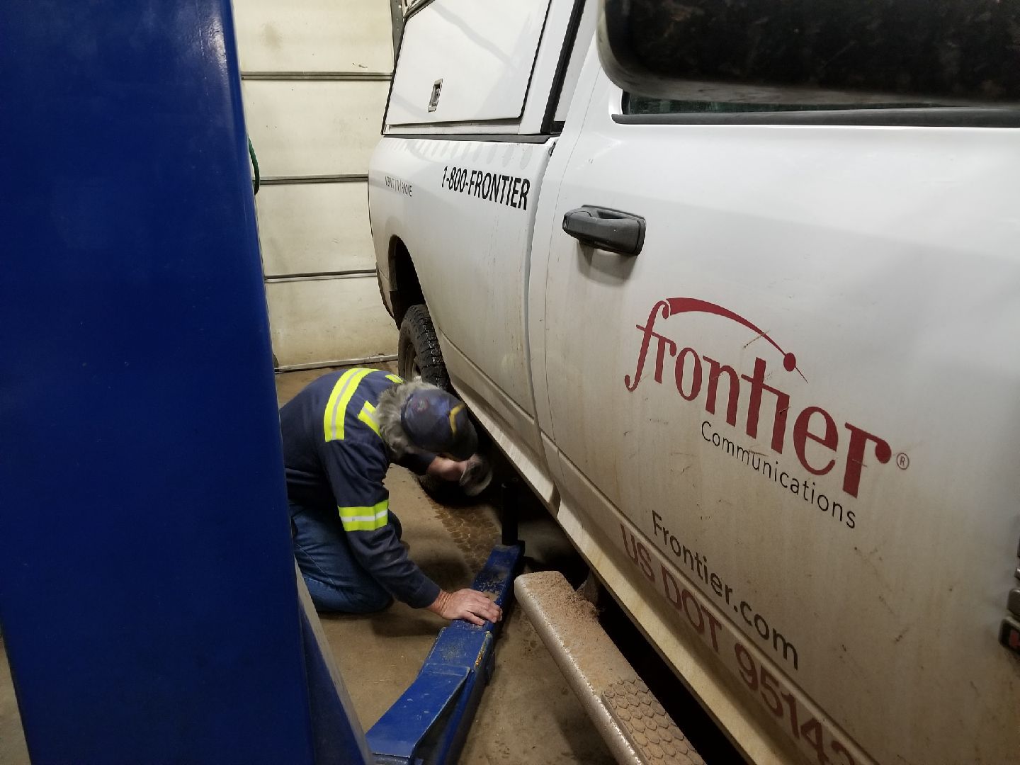 A technician in a high-visibility uniform kneels to position a blue hydraulic lift arm under a Frontier service truck.
