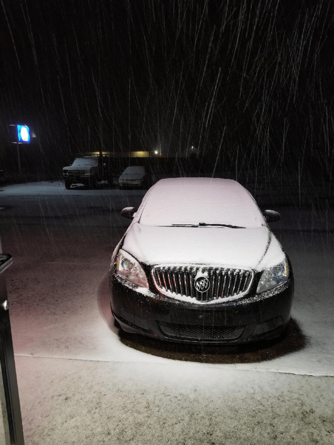 A black sedan covered in a layer of snow parked outside at night during a snowfall.