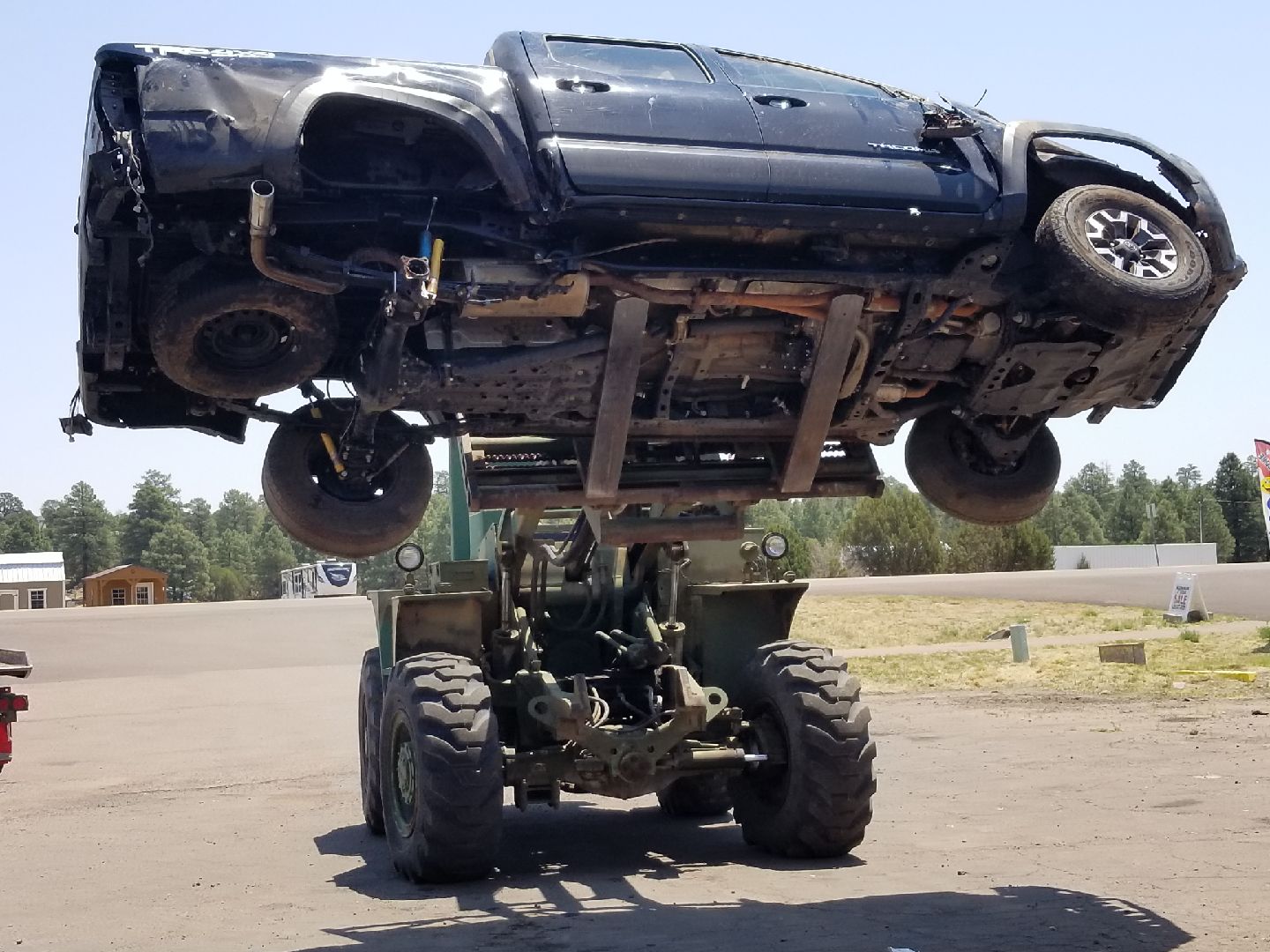 A large industrial forklift lifting a damaged dark pickup truck in an outdoor lot.