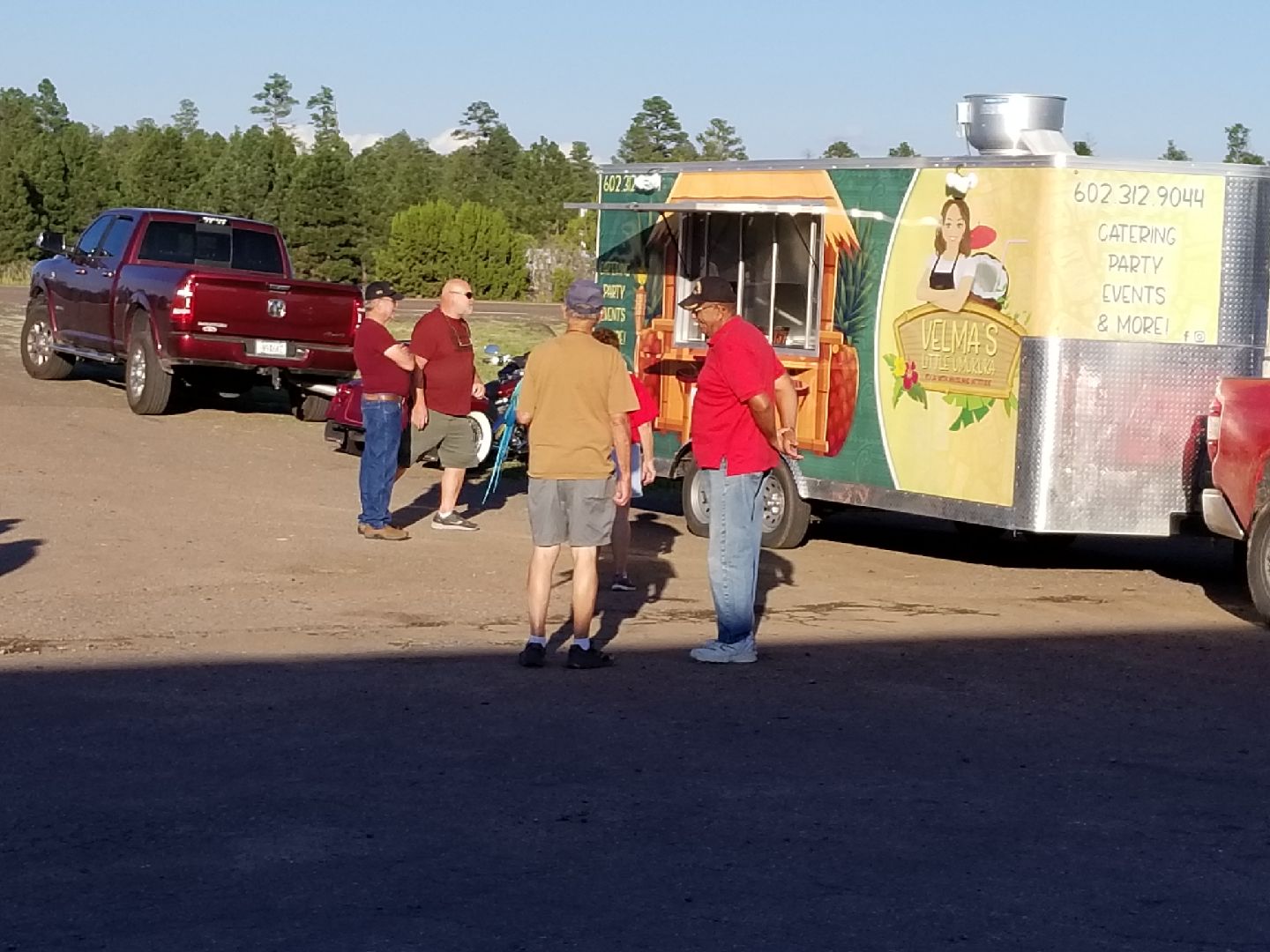 Four people stand near a food trailer in an outdoor parking lot next to a red pickup truck.