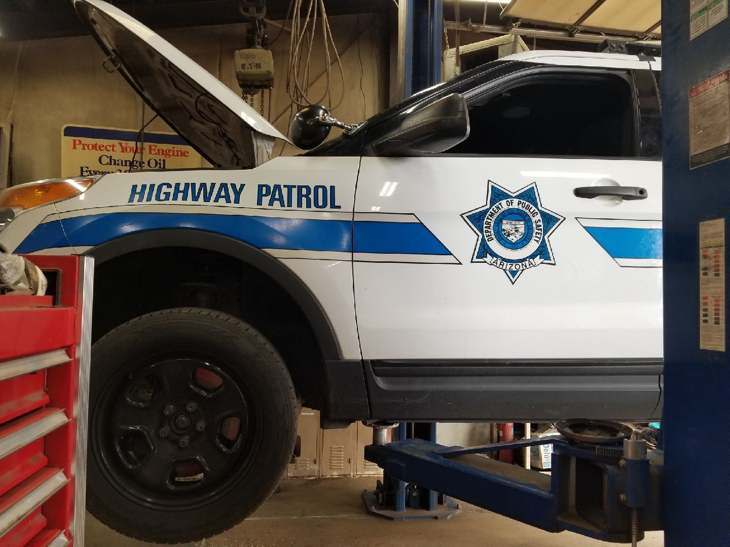 A white Highway Patrol vehicle with a blue stripe and star emblem is lifted on a mechanical hoist inside a garage.