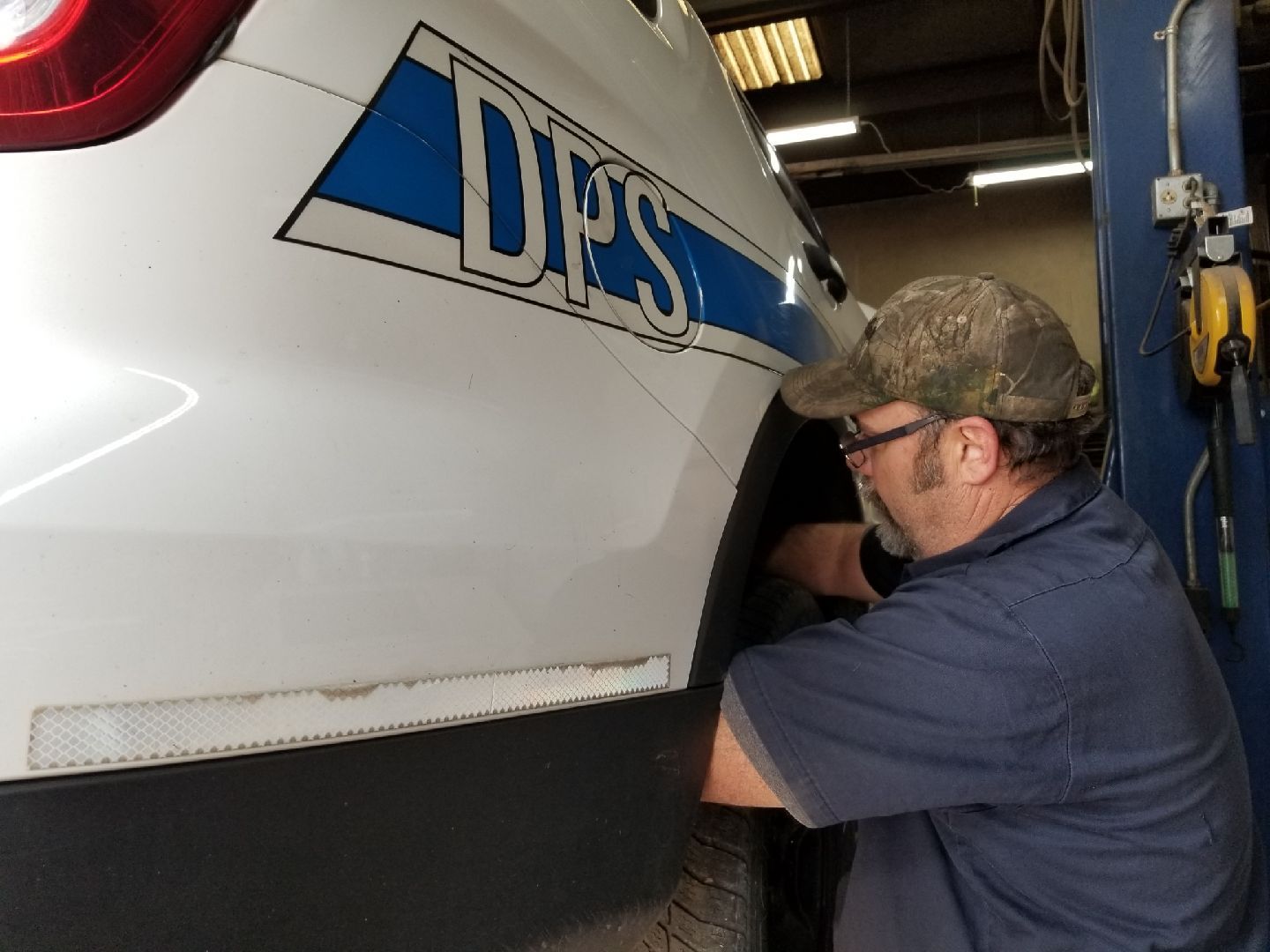 A mechanic wearing a camouflage hat and blue shirt works on the rear wheel of a white DPS police vehicle in a garage.