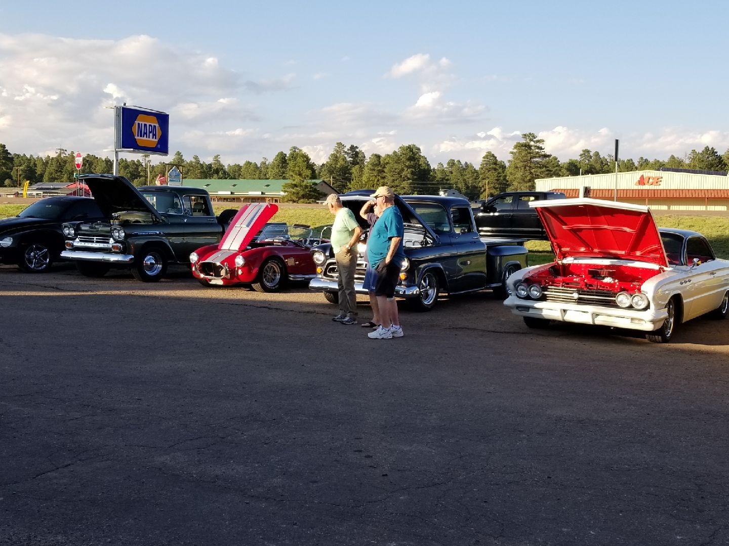 A row of classic cars with open hoods parked in an outdoor lot on a sunny day with two people standing nearby.