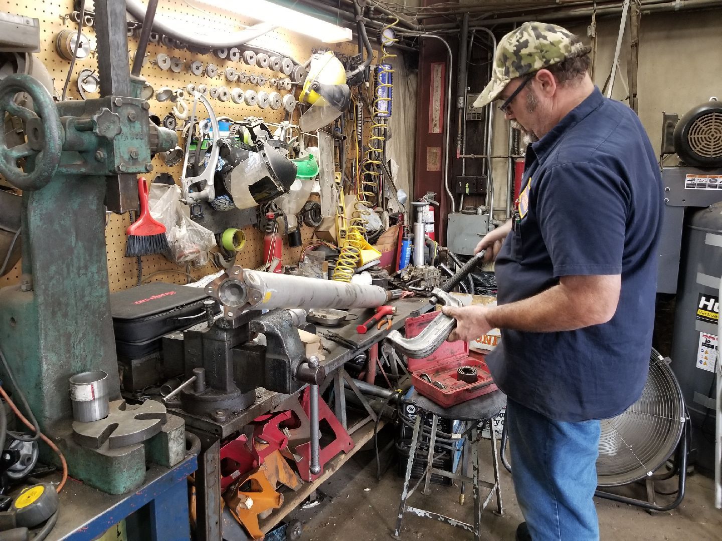 A mechanic wearing a camouflage cap works on a metal drive shaft in a cluttered automotive workshop.