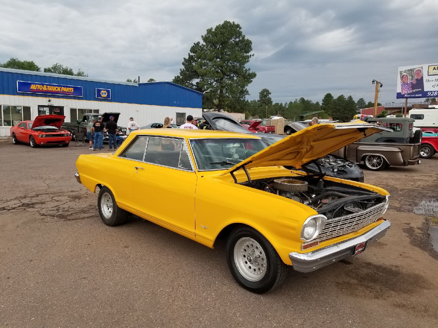 A bright yellow classic car with its hood open at an outdoor car show on a cloudy day.