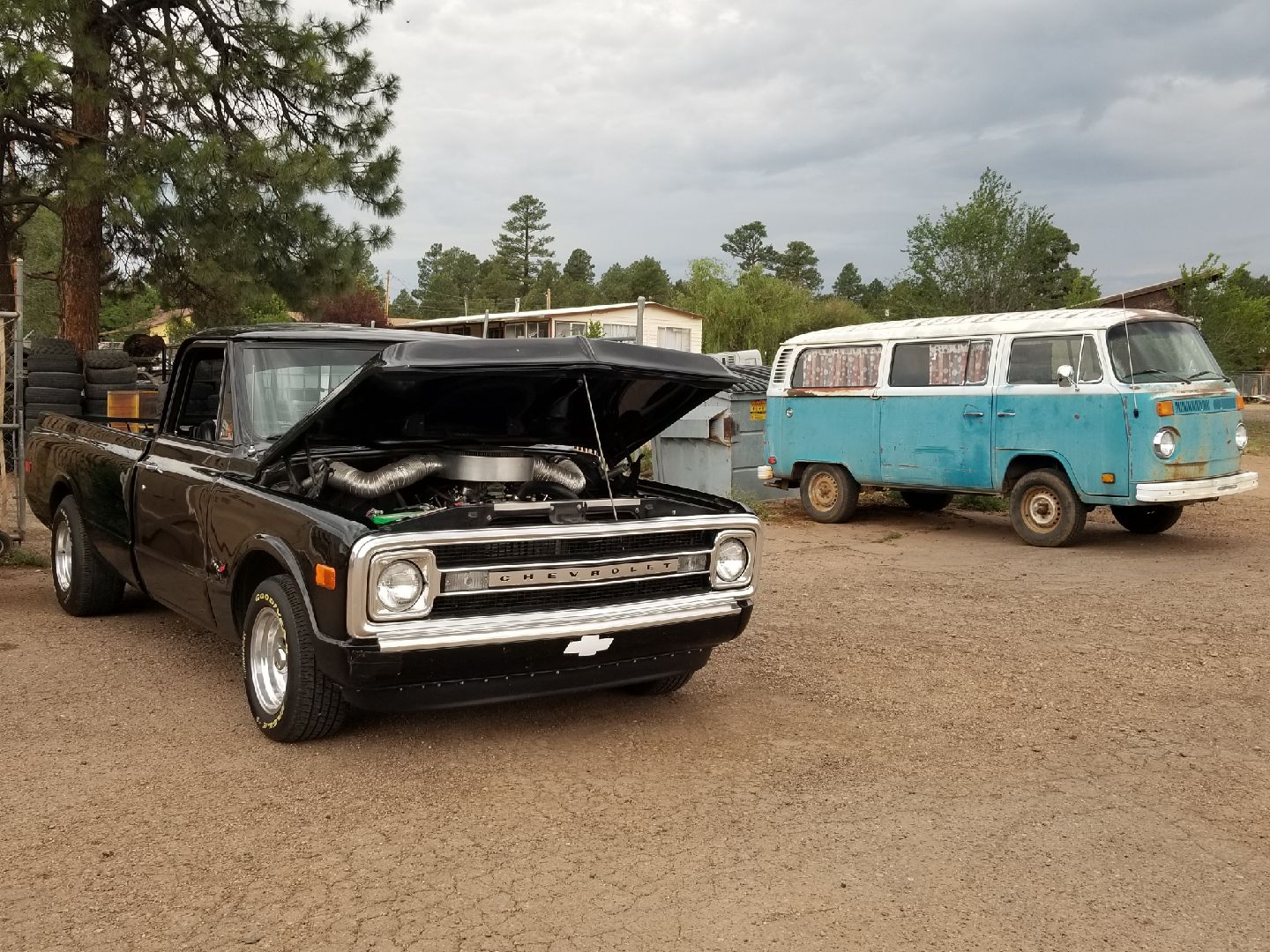 A black Chevrolet truck with its hood open and a vintage two-tone blue and white Volkswagen van on a gravel lot.