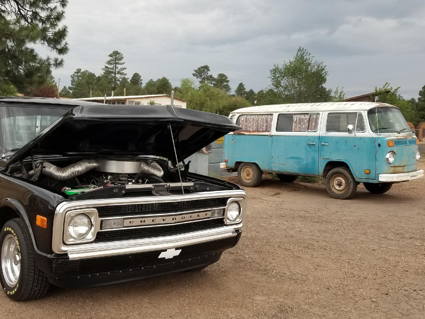 A black Chevrolet pickup with an open hood parked on gravel, with a vintage blue and white Volkswagen bus in the back.