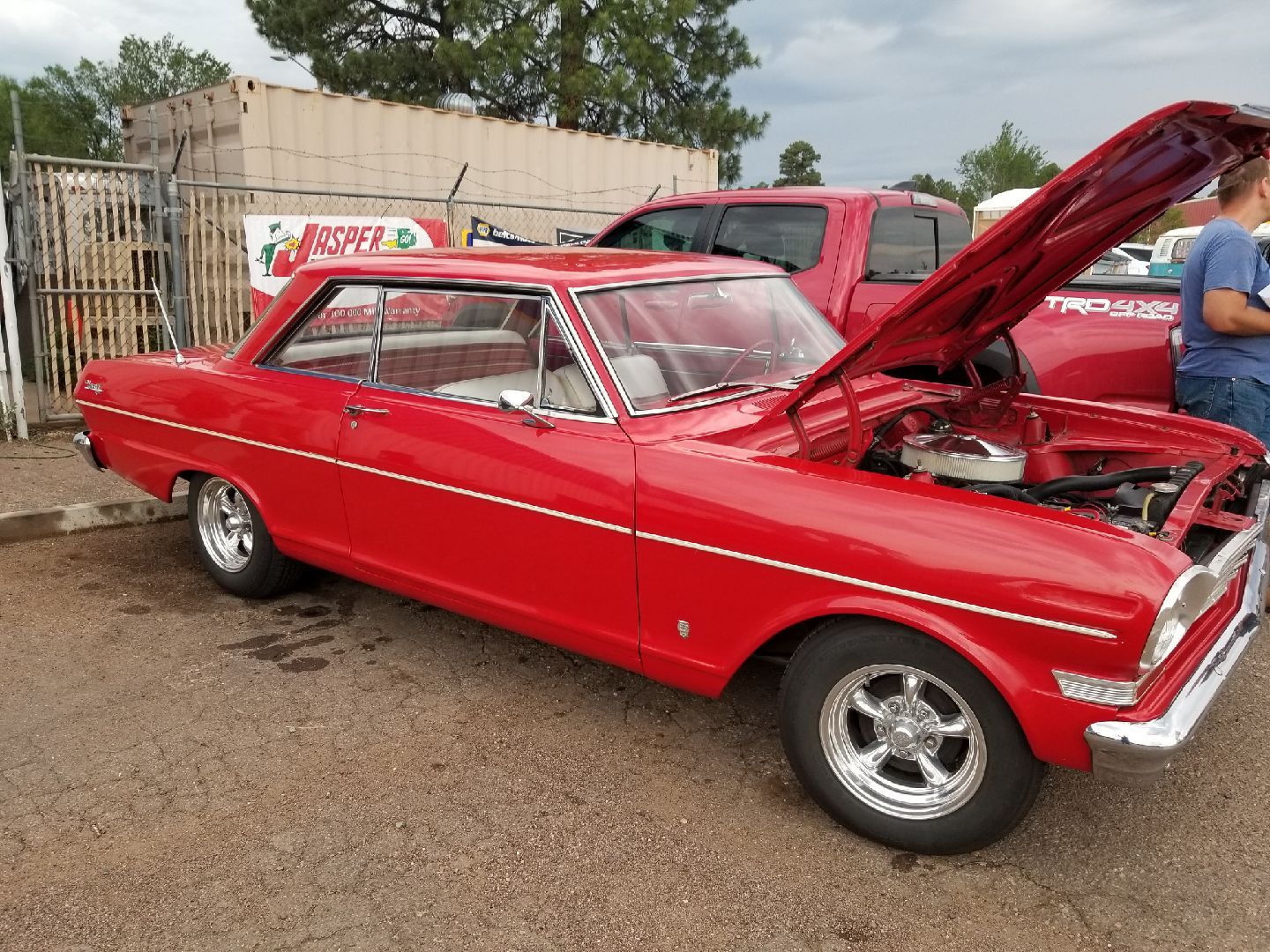 A bright red, vintage two-door Chevy Nova with its hood open at an outdoor car show.