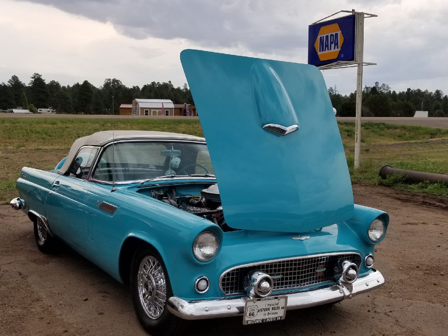 A turquoise vintage Ford Thunderbird convertible with its hood open, parked in a dirt lot beneath an NAPA Auto Parts sign.
