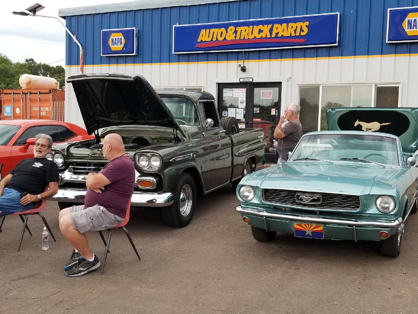 Two people sit in chairs in front of a NAPA auto parts store near a vintage dark green truck and a light blue Mustang.