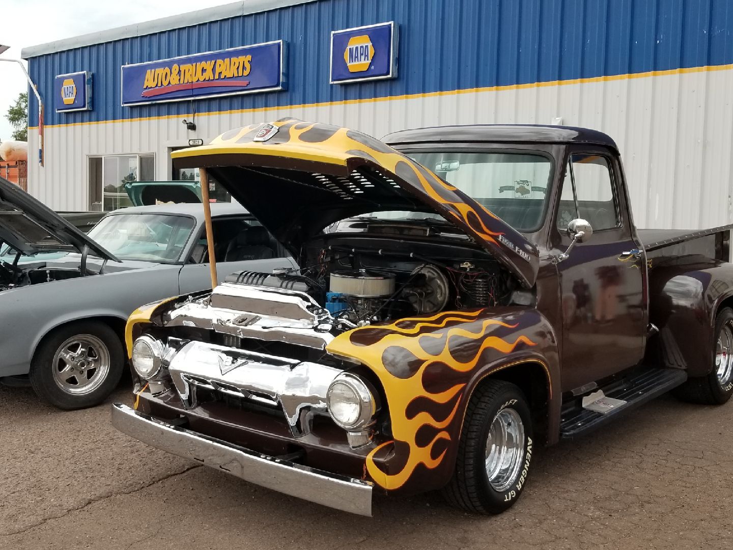Brown vintage pickup truck with yellow flames, hood open, parked in front of a NAPA Auto Parts store.