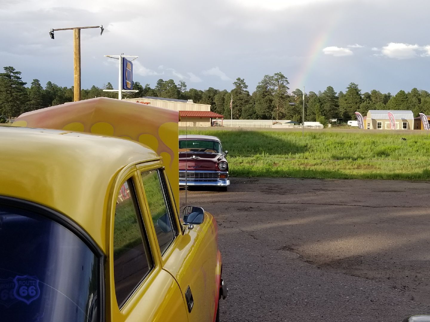 A yellow vintage car with its hood up faces a maroon classic car in a gravel lot with a faint rainbow in the sky above.