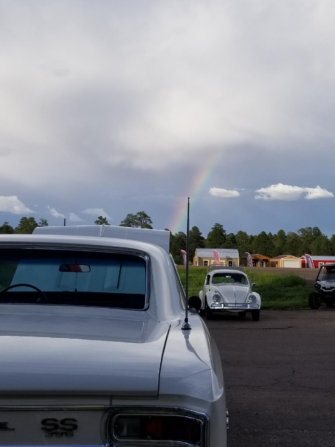 A silver vintage car and a white VW Beetle parked in a lot under a cloudy sky with a faint rainbow.