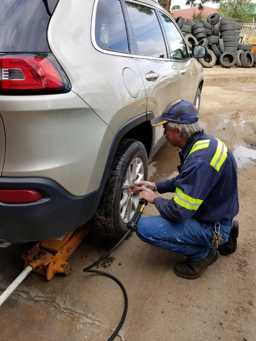 A mechanic in high-visibility work gear uses a pneumatic impact wrench to change the tire of a beige SUV outdoors.