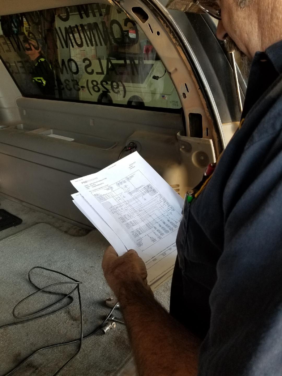A person in a dark blue uniform holds a technical wiring diagram inside the rear of a vehicle with an open side panel.