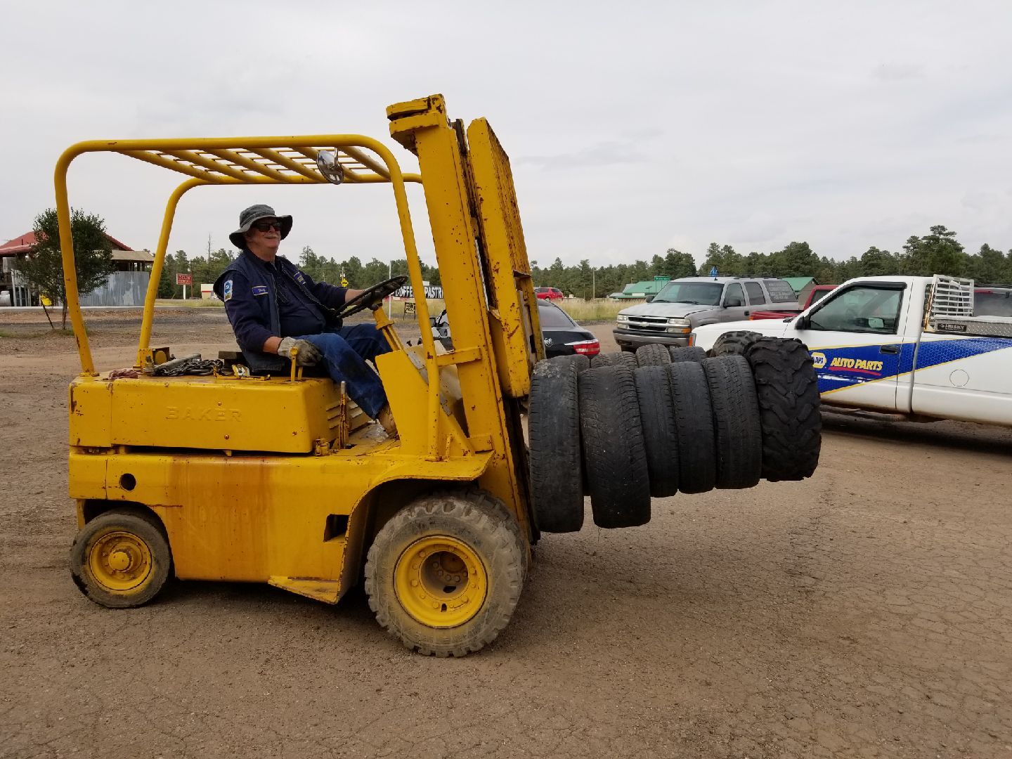 A person operates a yellow forklift carrying a stack of large tires on a dirt lot with vehicles parked in the background.