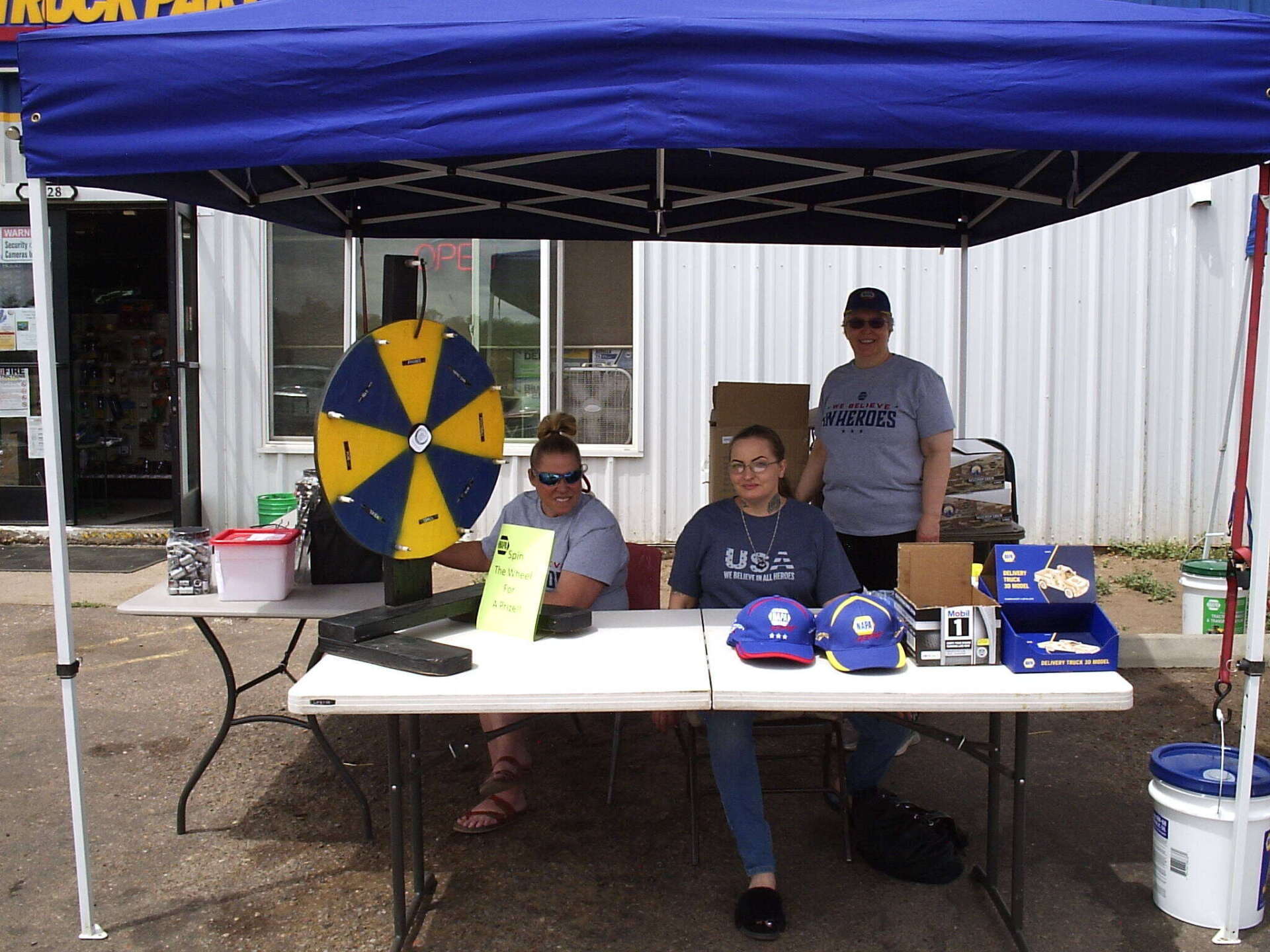 Three people sit at a table under a blue canopy with a prize wheel and promotional items outside a building.
