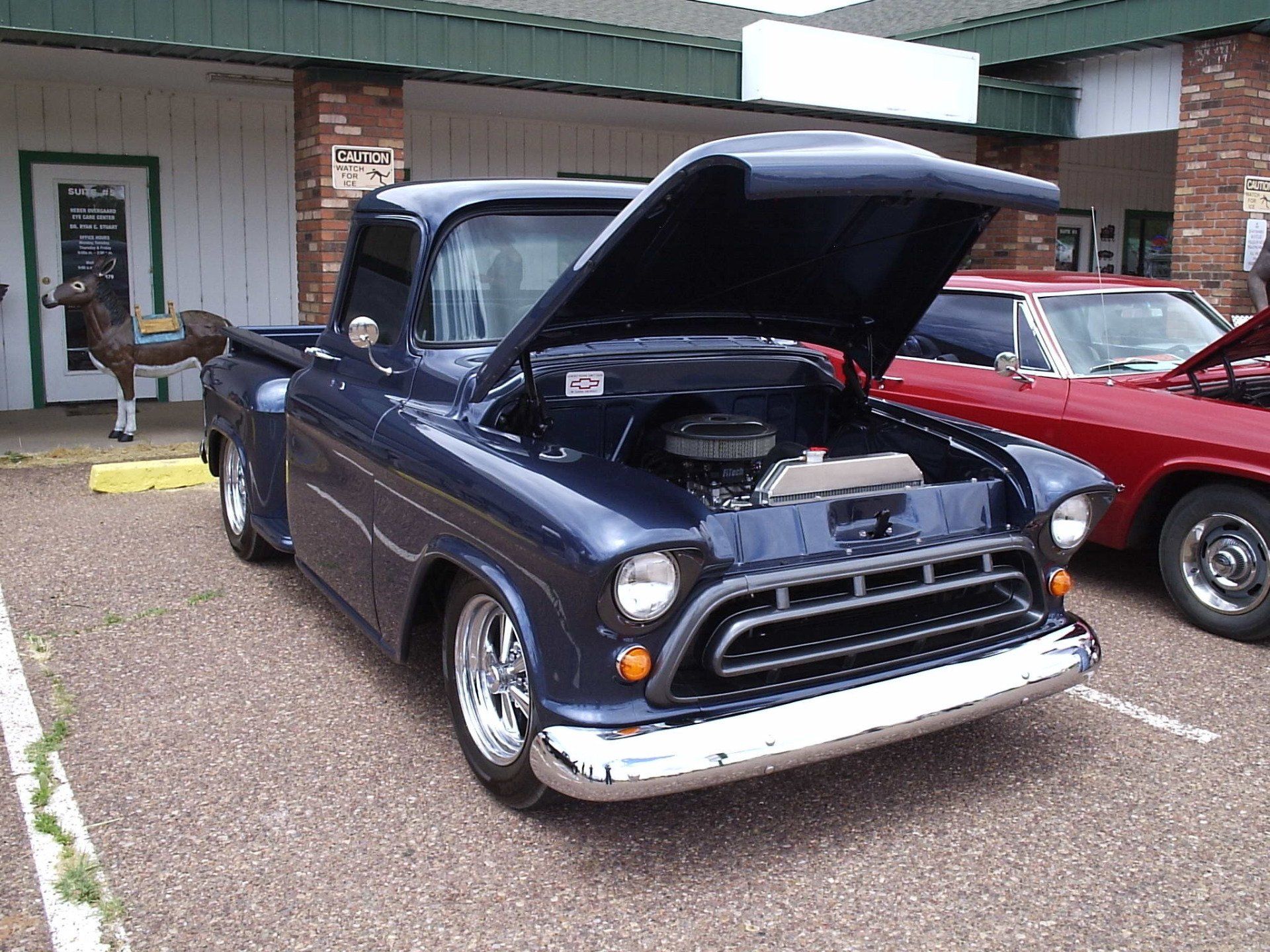 A dark blue vintage Chevrolet truck with its hood open, parked in a parking lot next to a red car.