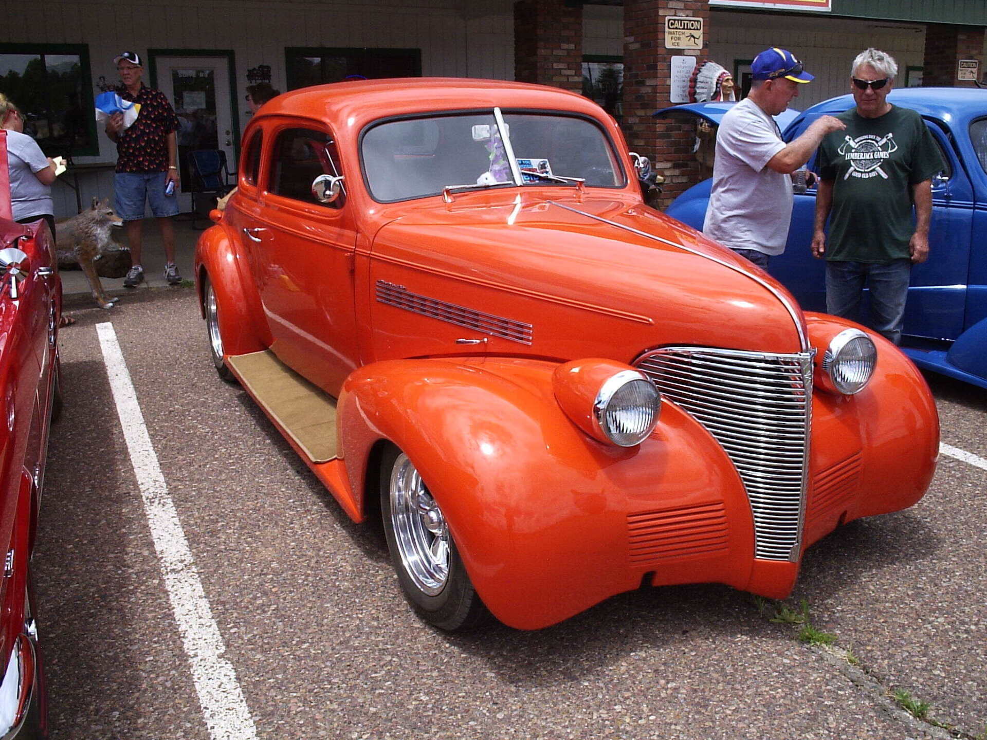 A bright orange vintage hot rod car parked outdoors, with two people standing nearby and a blue car to the right.