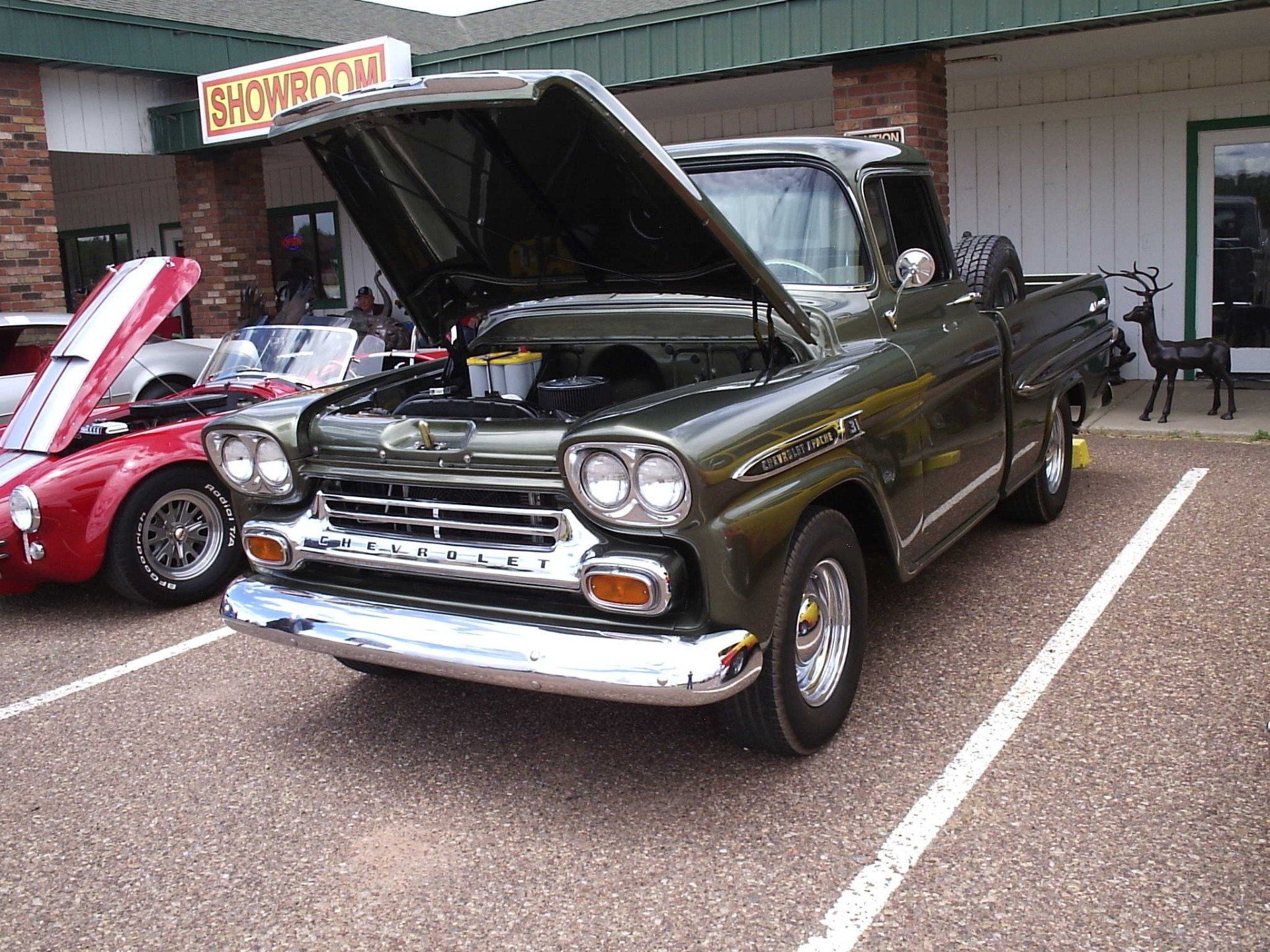 An olive green vintage Chevrolet pickup truck with its hood open, parked in a gravel lot next to a red sports car.