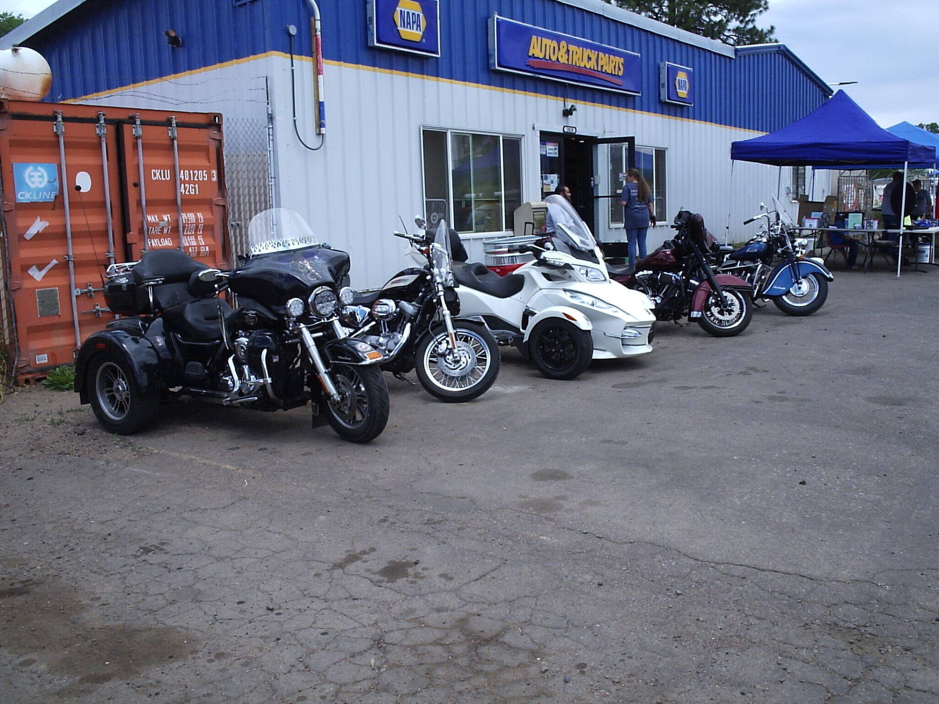 A row of motorcycles and a three-wheeled motorcycle parked in front of a blue and white building with a blue canopy.