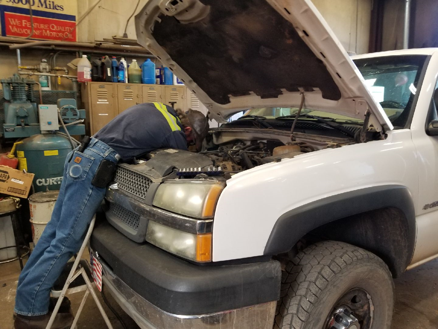 A technician standing on a step ladder working on the engine of a white pickup truck inside an auto repair shop.