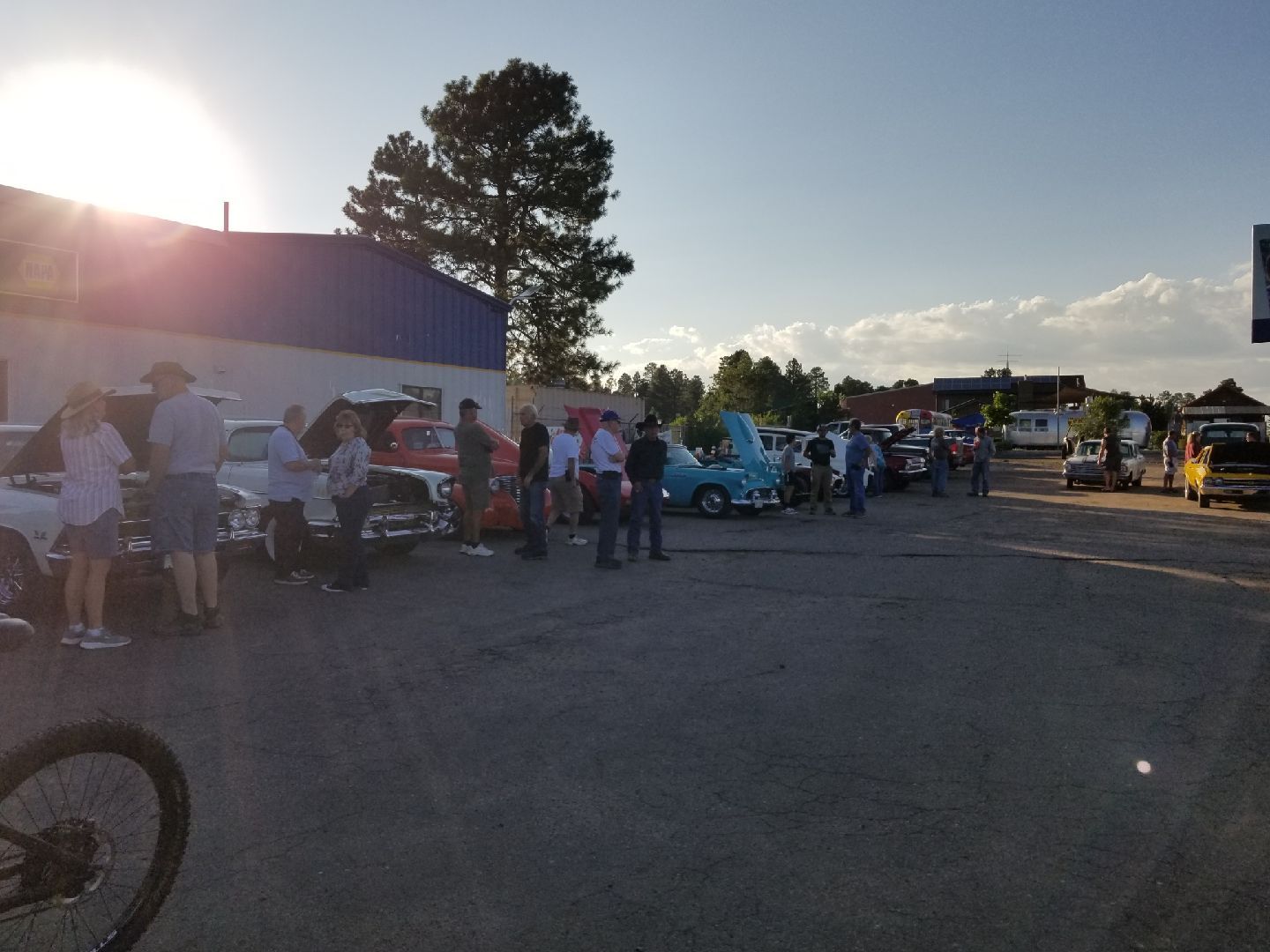 People gather in an outdoor parking lot at sunset to look at classic cars with open hoods.