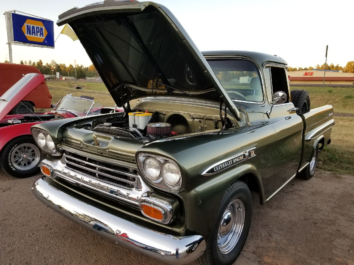 A dark green vintage Chevrolet Apache pickup truck with its hood open at an outdoor car show near a NAPA sign.
