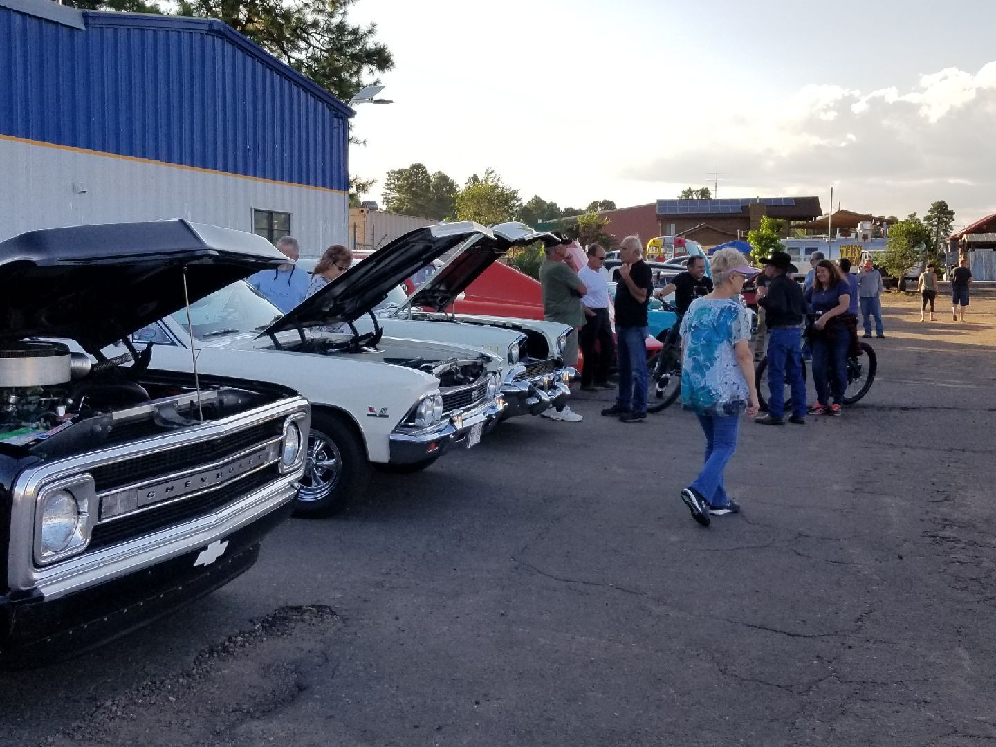 People gather at an outdoor car show in a parking lot, viewing classic vehicles with their hoods raised.