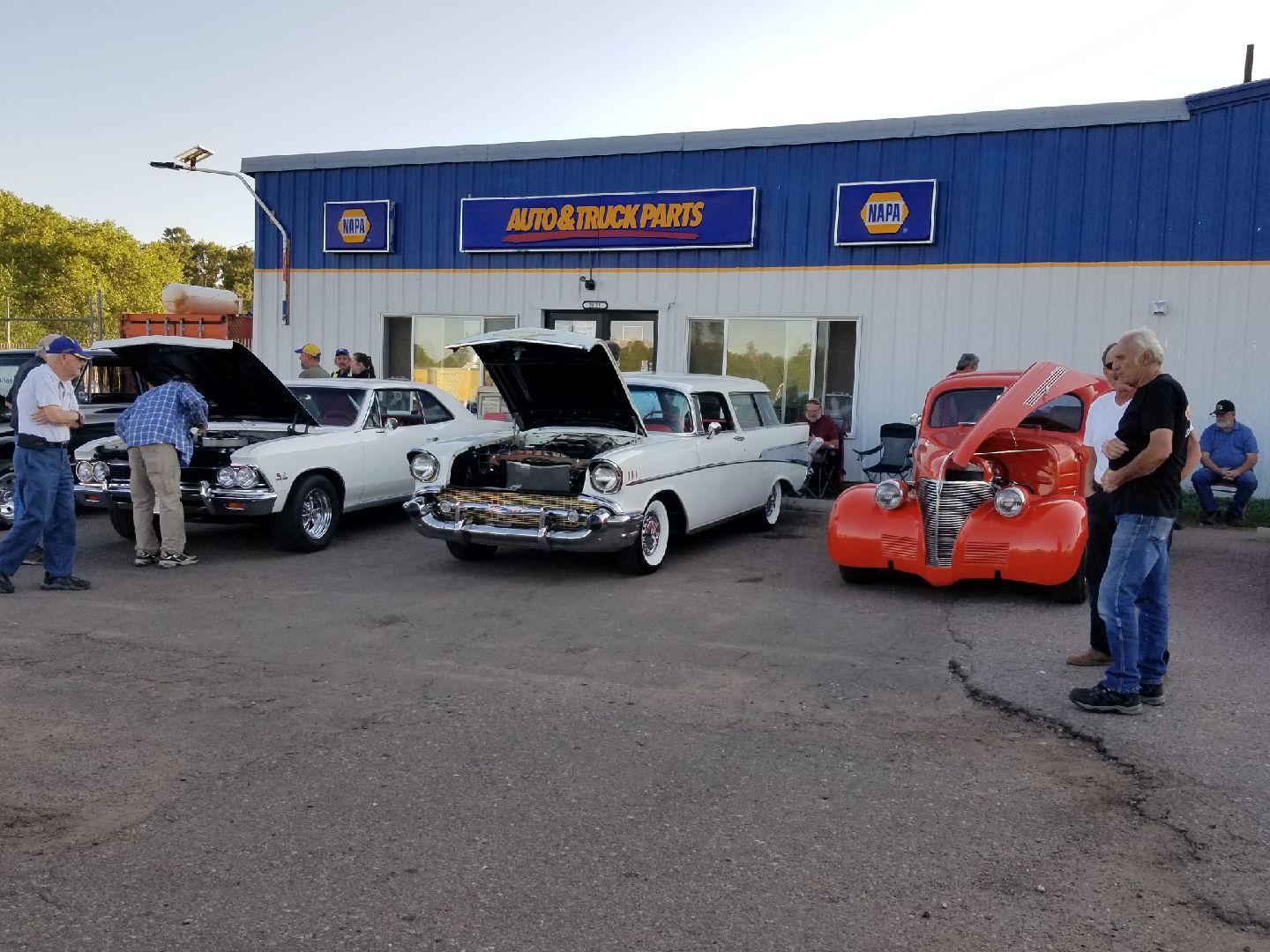 Classic cars on display at a NAPA Auto & Truck Parts store, with people standing nearby looking at the vehicles.