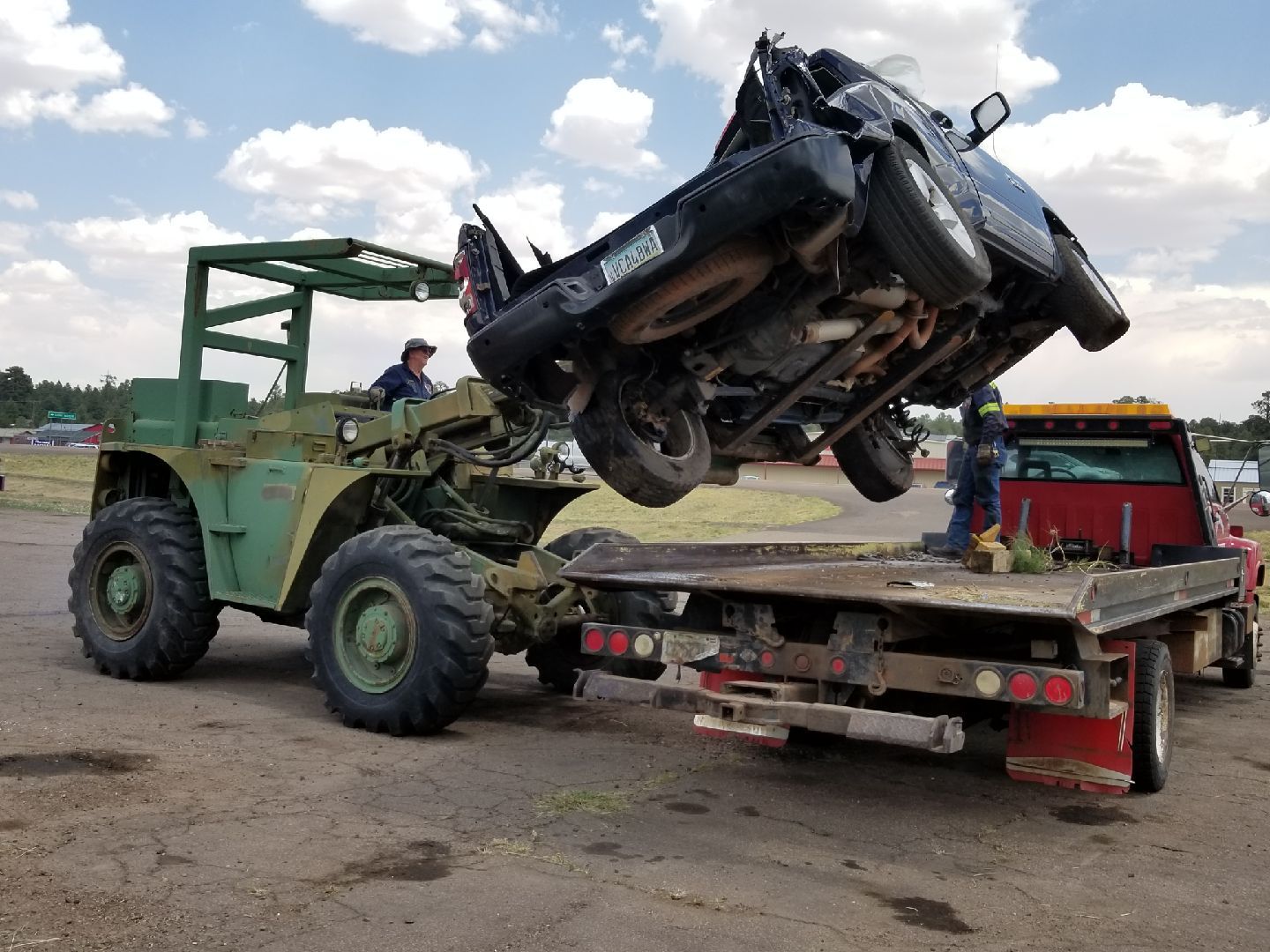 A green forklift lifts a damaged black vehicle onto a flatbed truck outdoors under a cloudy sky.