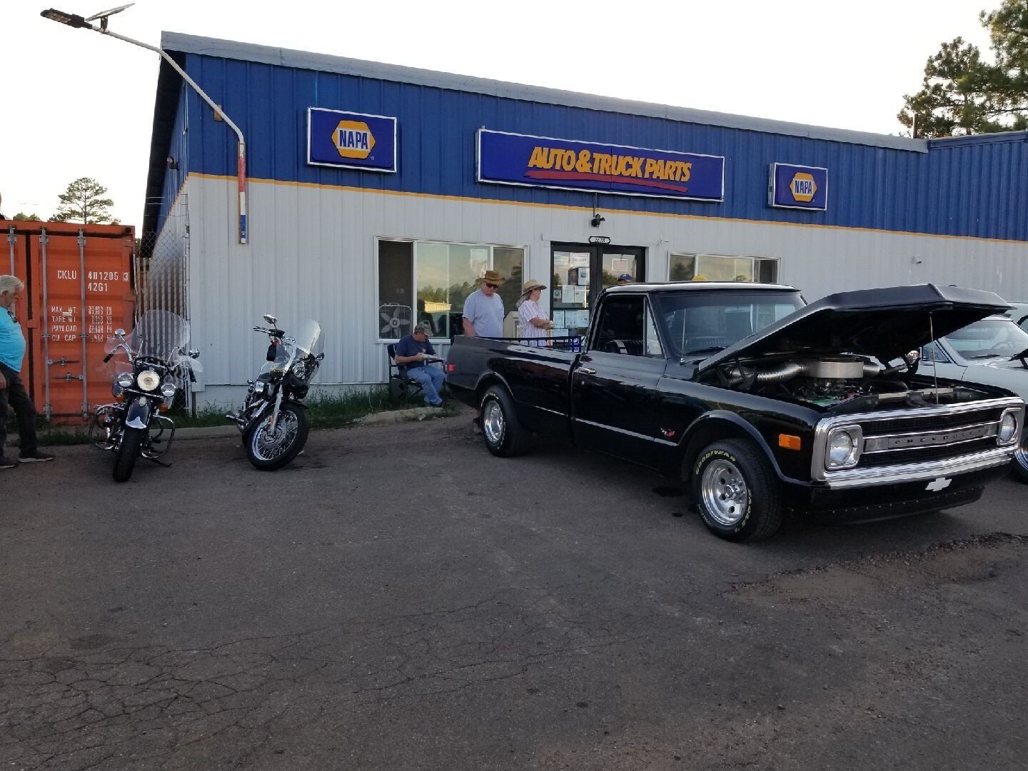 A black vintage pickup truck with an open hood parked outside an Auto & Truck Parts store with motorcycles nearby.