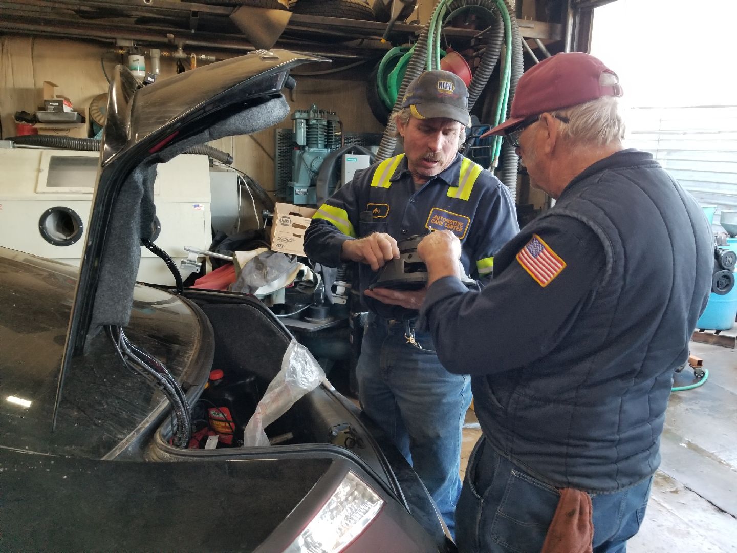 Two workers in a workshop inspect an item inside the open trunk of a car.